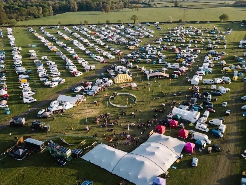 Aerial photo of a campervan festival in a green field. Neat rows of various campervans and motorhomes and a communal area full of people.